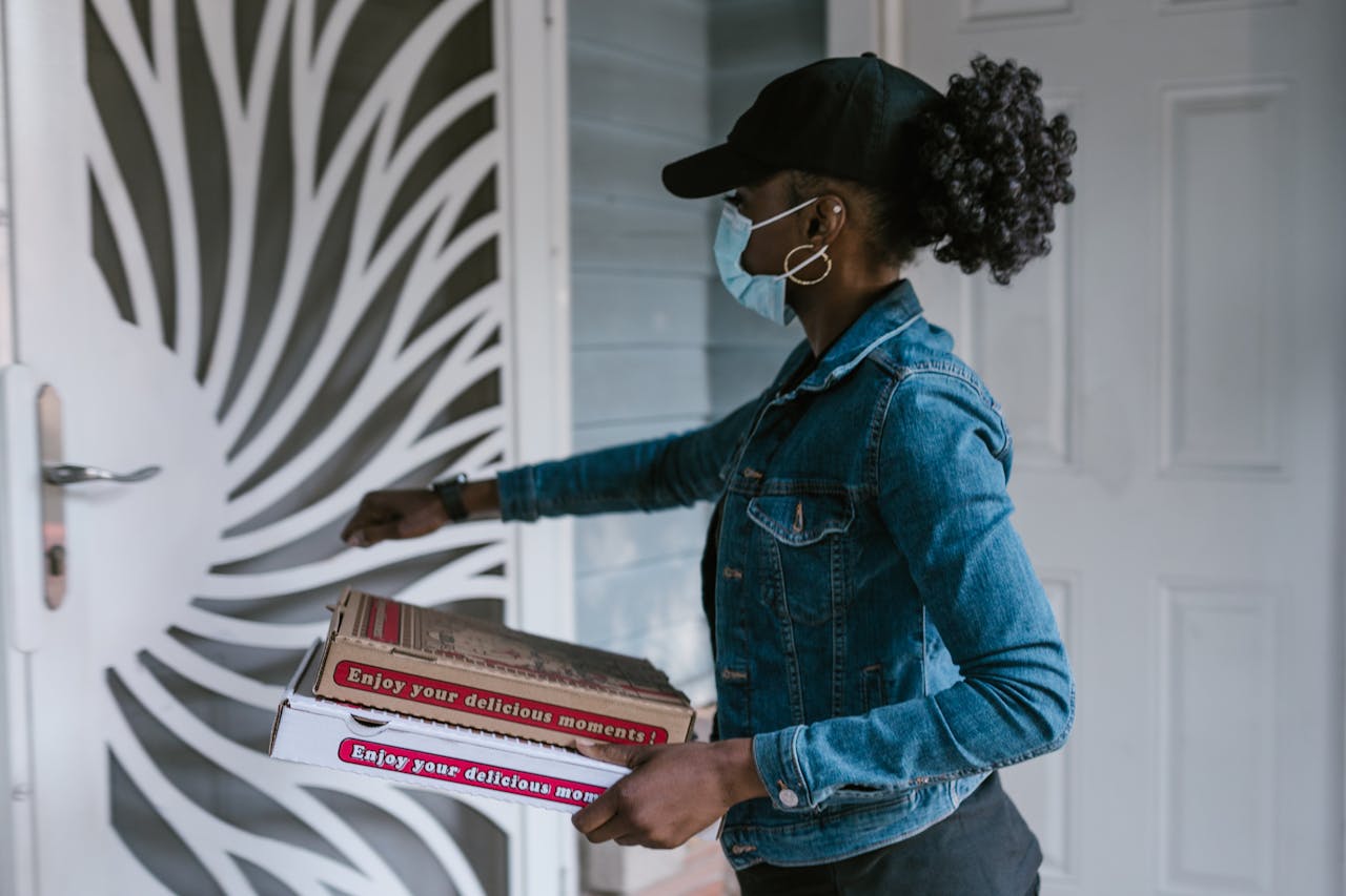 Black woman in denim jacket delivering pizza with a face mask at the door.